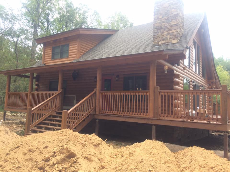 Wooden cabin with a wraparound porch and stone chimney, surrounded by trees and dirt in the foreground
