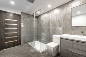 Modern bathroom with gray tiles, glass shower, vanity, mirror, and white toilet