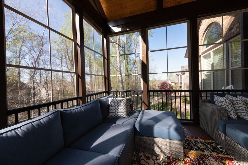 Sunlit screened porch with dark blue sectional sofas and a small ottoman, overlooking trees and a patio view