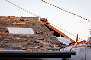 Damaged roof with missing shingles, exposed wood, and bent utility wires against a white sky