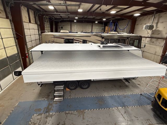 RV with white awning extended in a garage, next to other RVs. Black tires, blue floor.