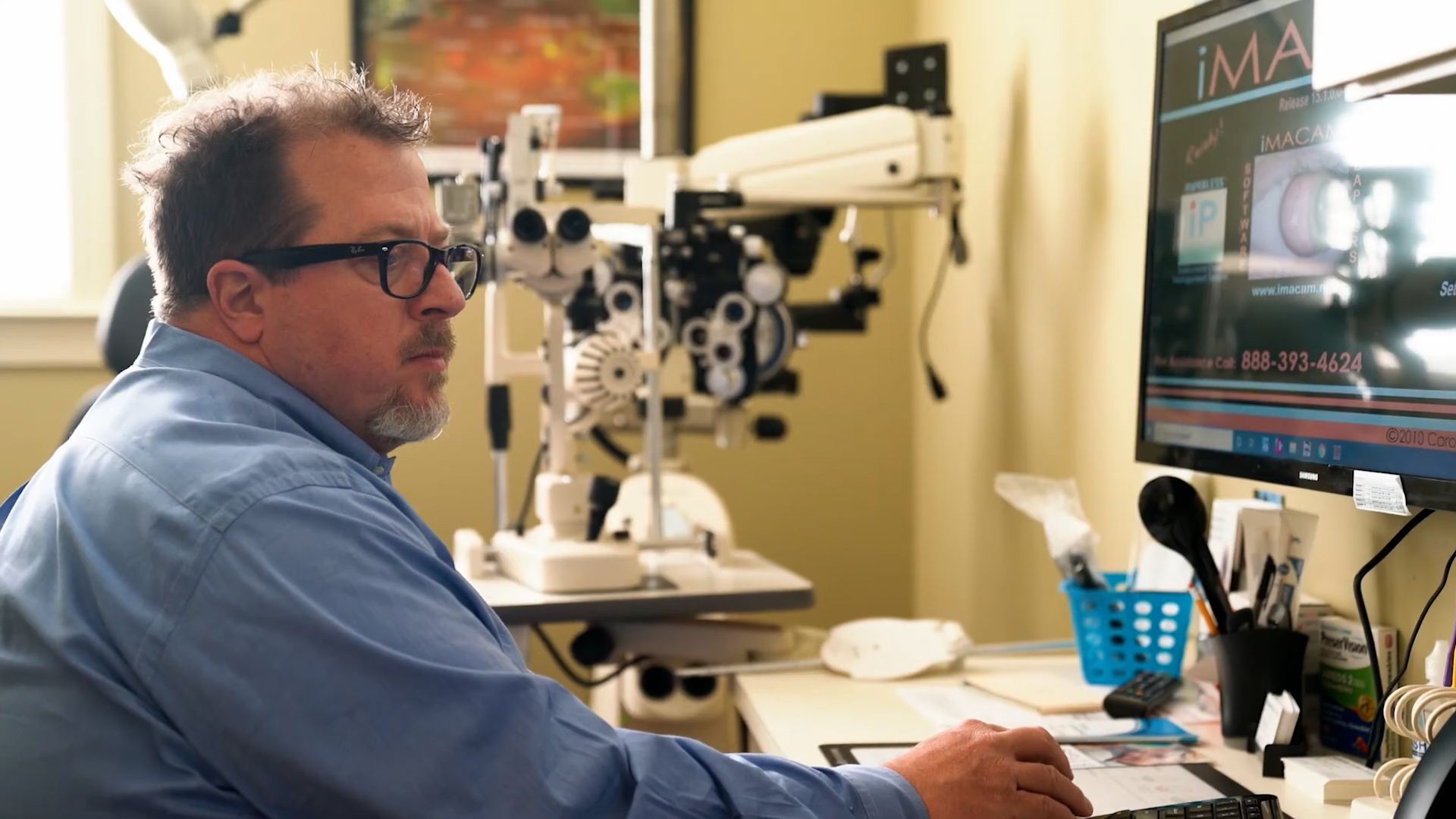 Man at a desk using a computer in a medical office, with an eye exam microscope beside him near Fayetteville, NC.