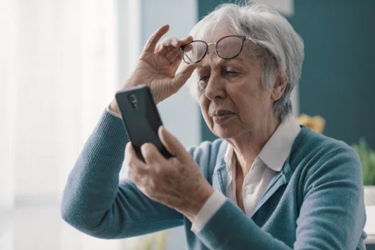 Senior person adjusting glasses while looking at a smartphone in a bright room near Fayetteville, NC.