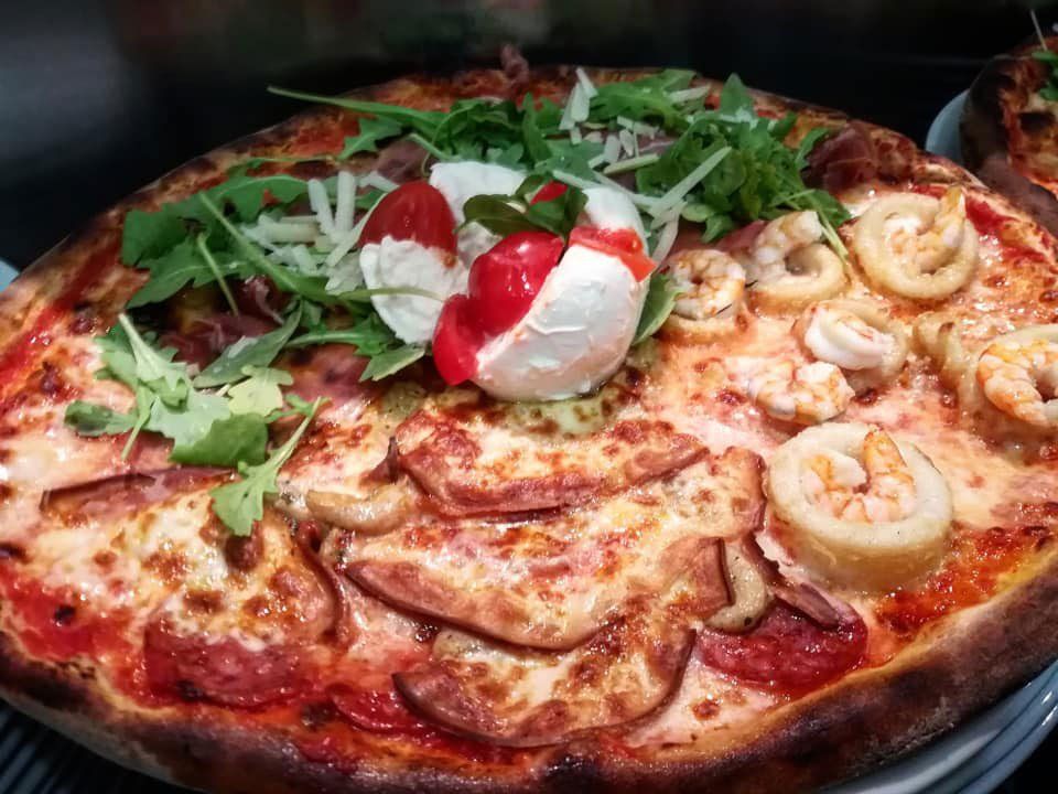 Una pizza condita con gamberi, salame, una grossa palla di burrata e rucola su uno sfondo scuro.