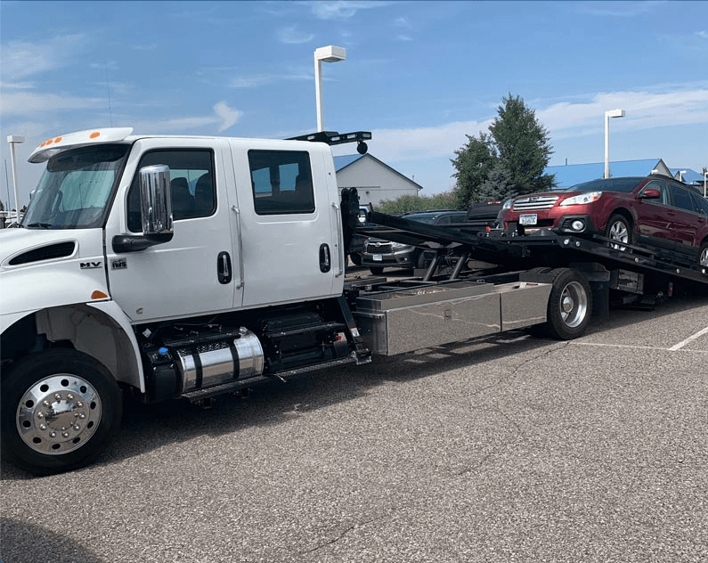 A tow truck is towing a red car in a parking lot.