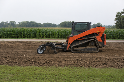 Orange compact tractor grading a dirt field with a cornfield in the background