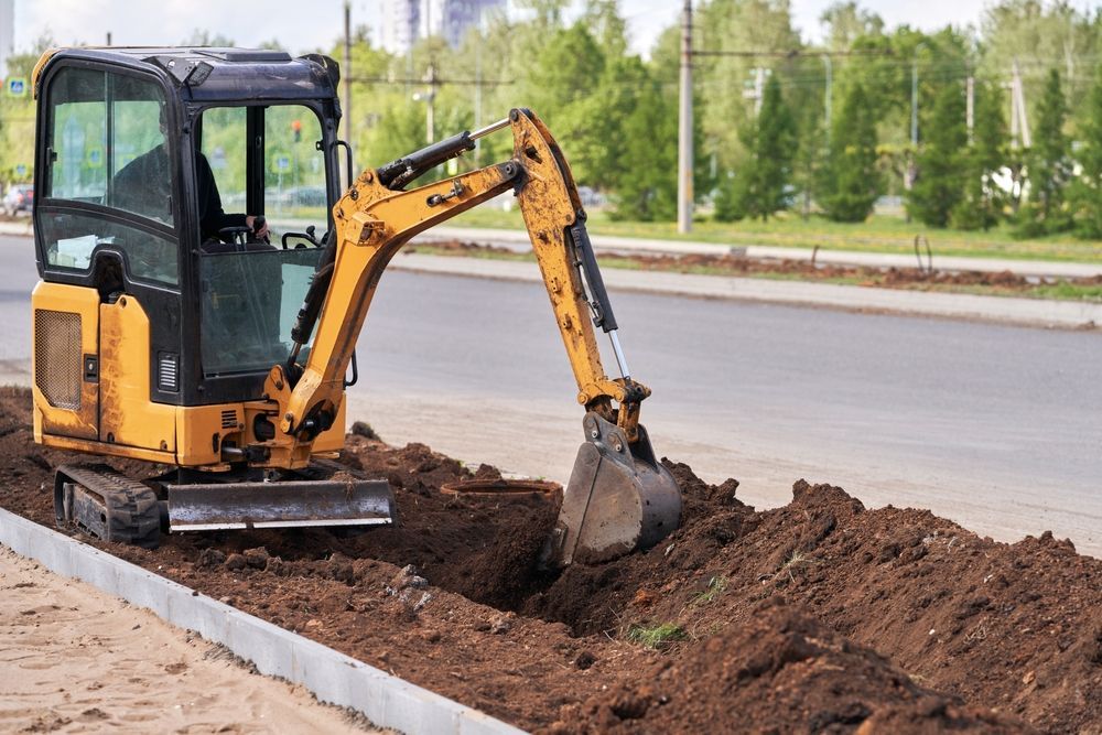 Yellow mini excavator digging a roadside trench beside a curb and road