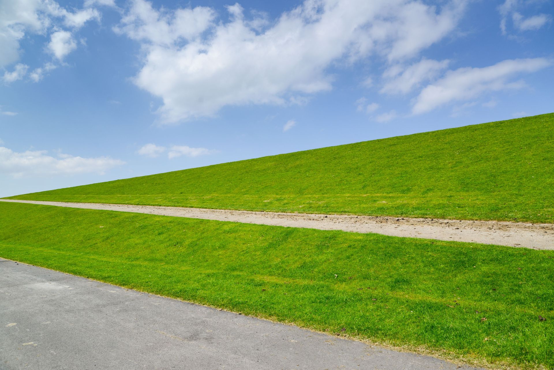 Grassy hill with a winding dirt path under a blue sky with clouds, beside a paved road