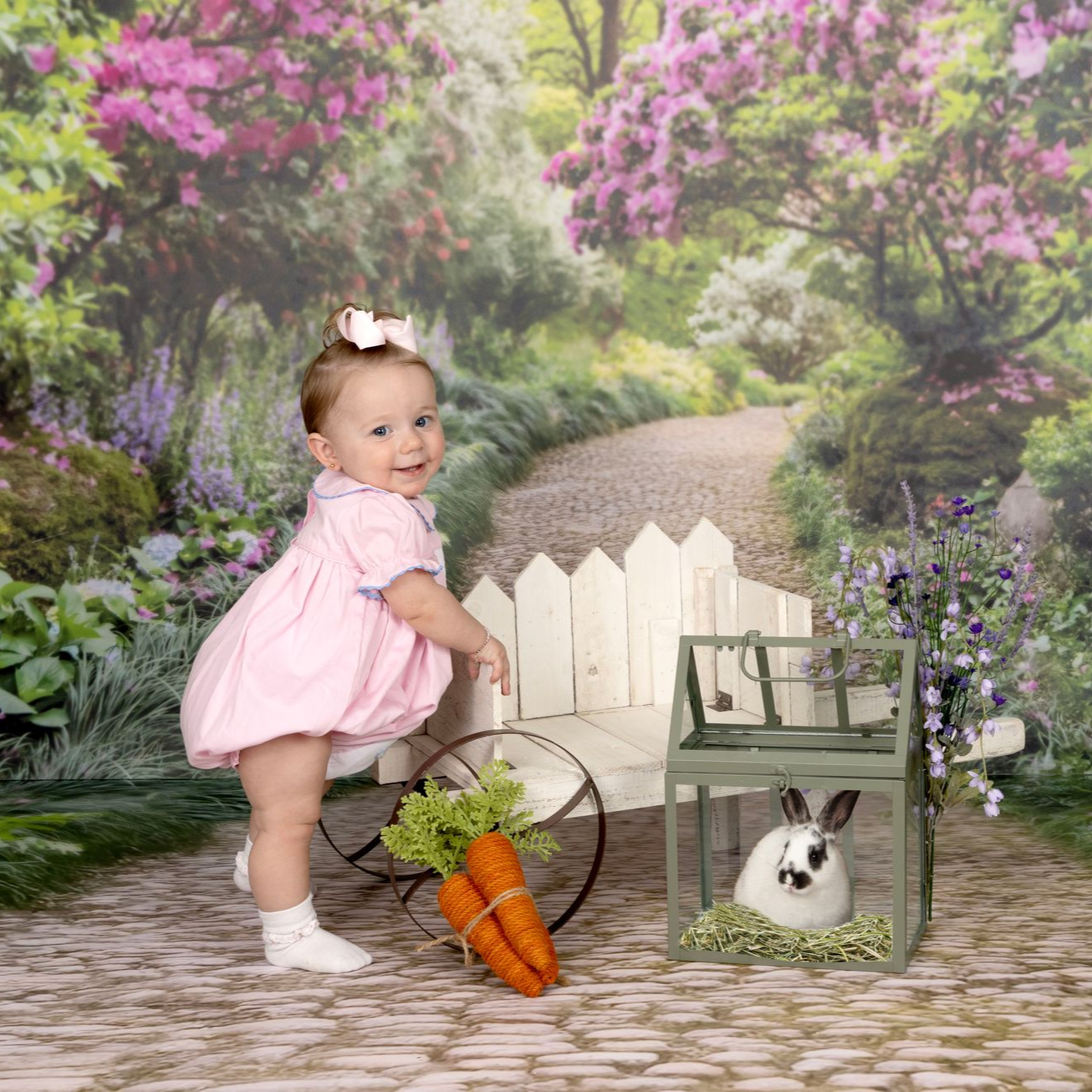 Baby in pink dress by a white fence in a garden scene with a rabbit and carrots in a basket