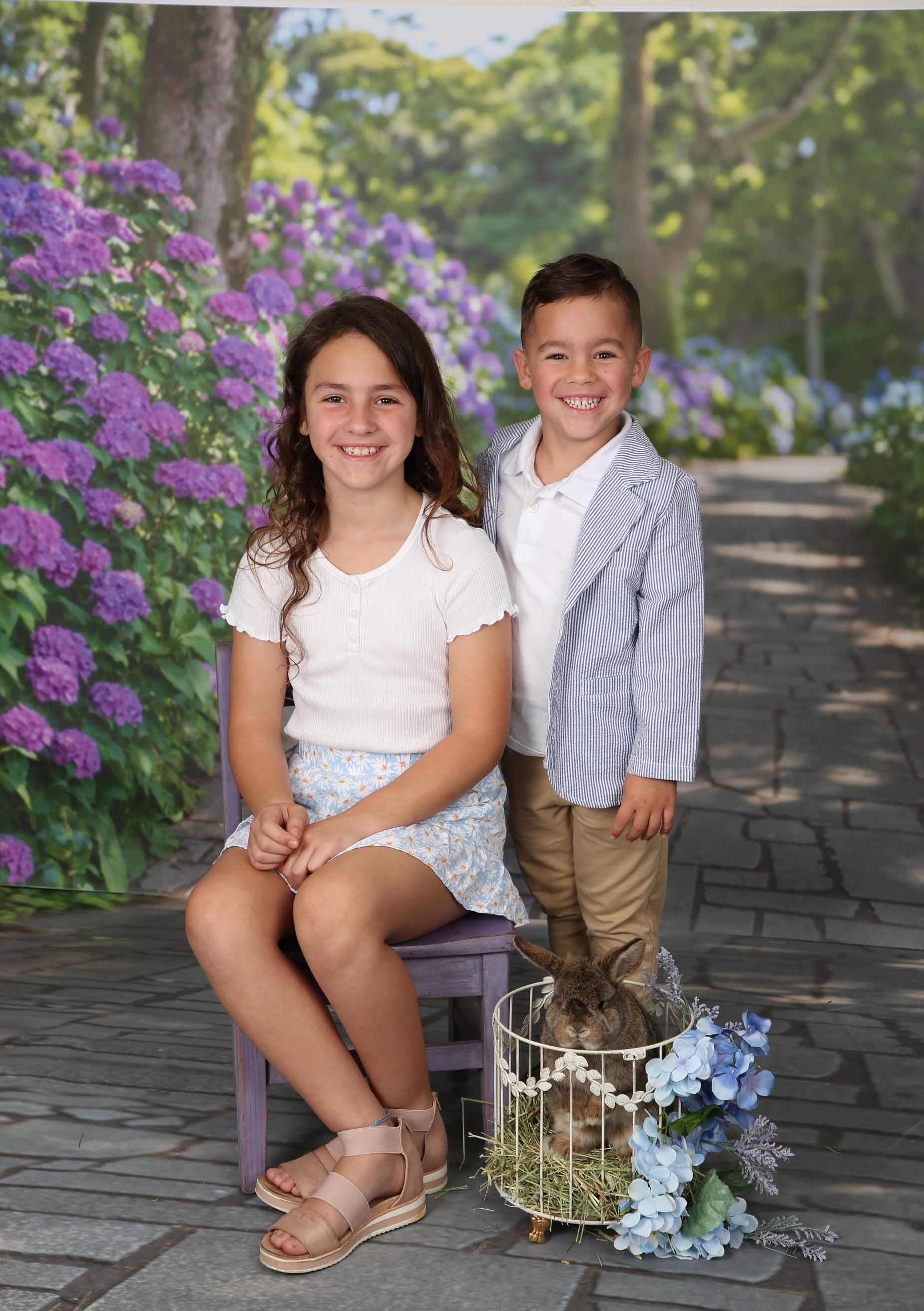 Two children posing on a stone path beside purple flowers, with a basket of blue blooms nearby