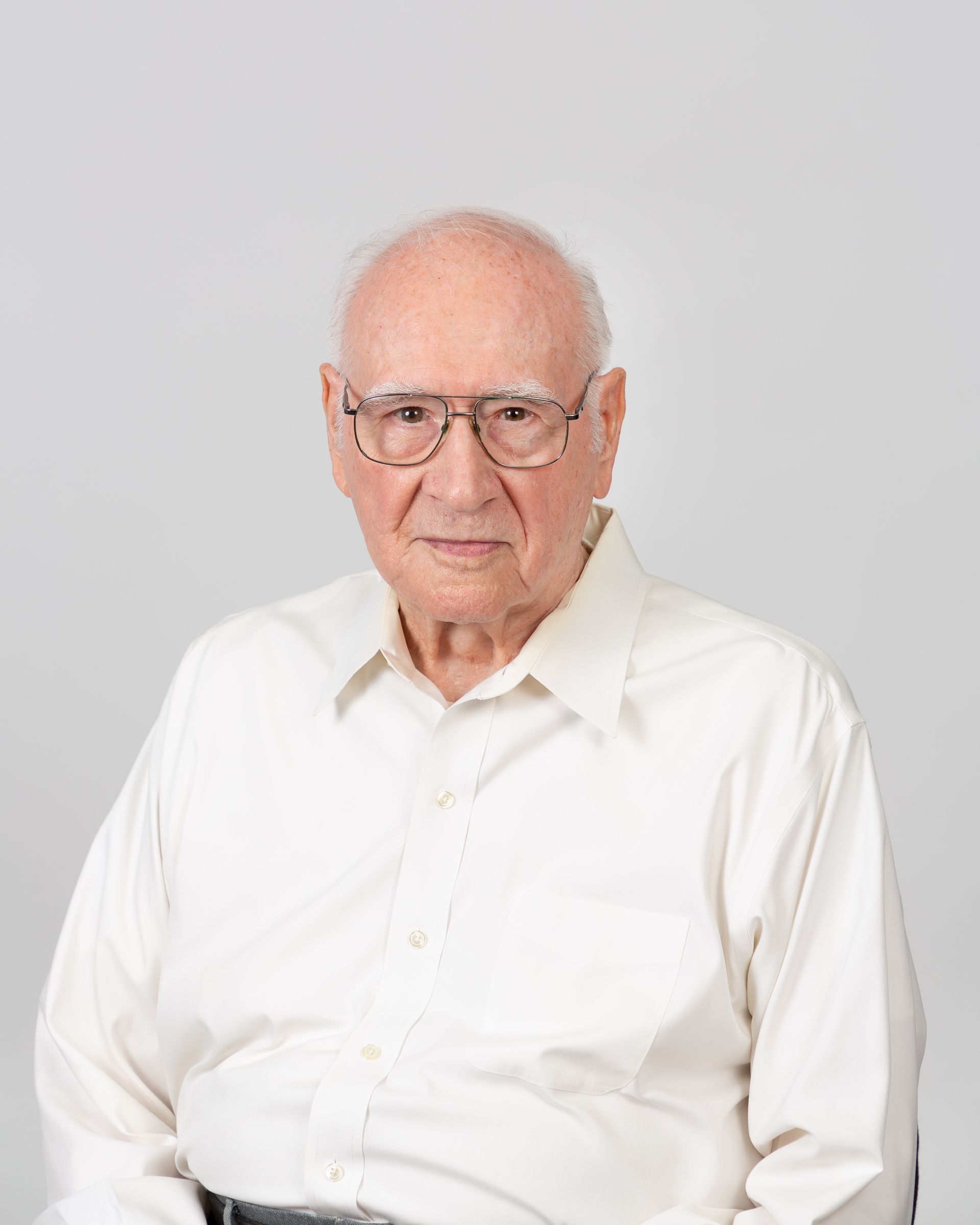 Elderly man in a white shirt and glasses, seated against a light gray background.
