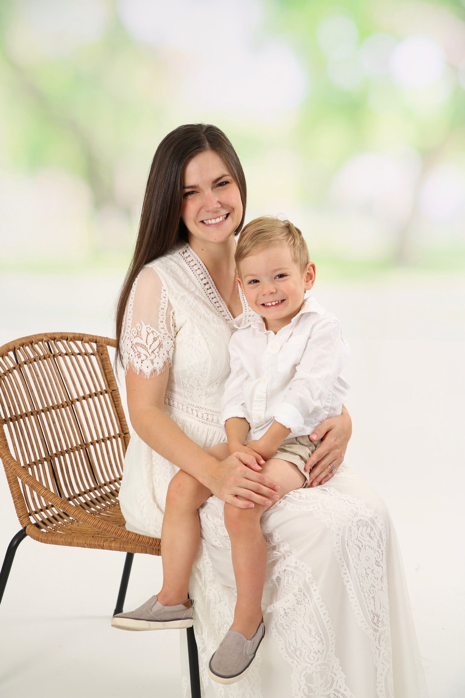 Woman in white dress sitting with a smiling child on her lap beside a wicker chair, against a soft green background