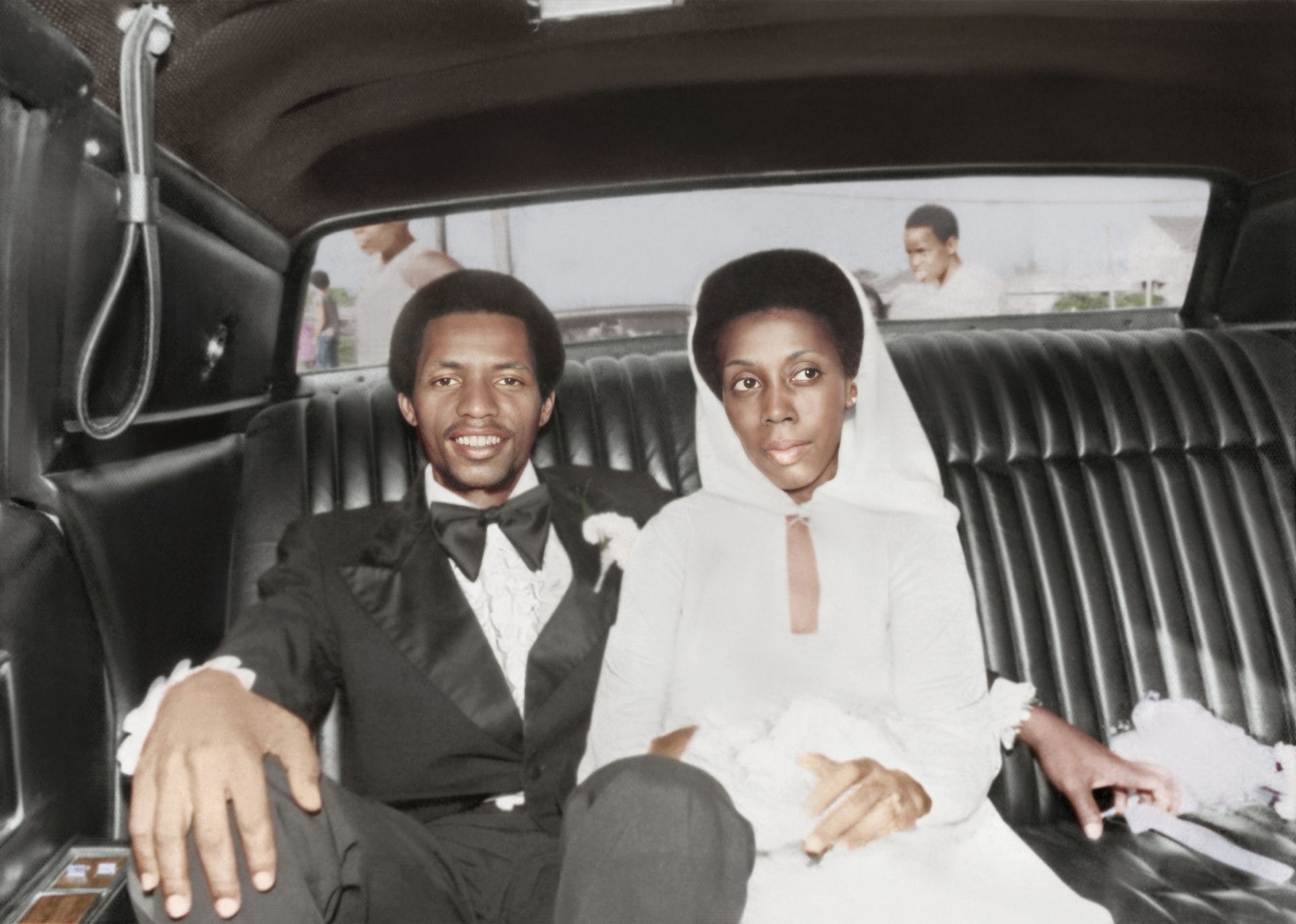 Couple seated in a car’s back seat, dressed formally, with a city street visible behind them.