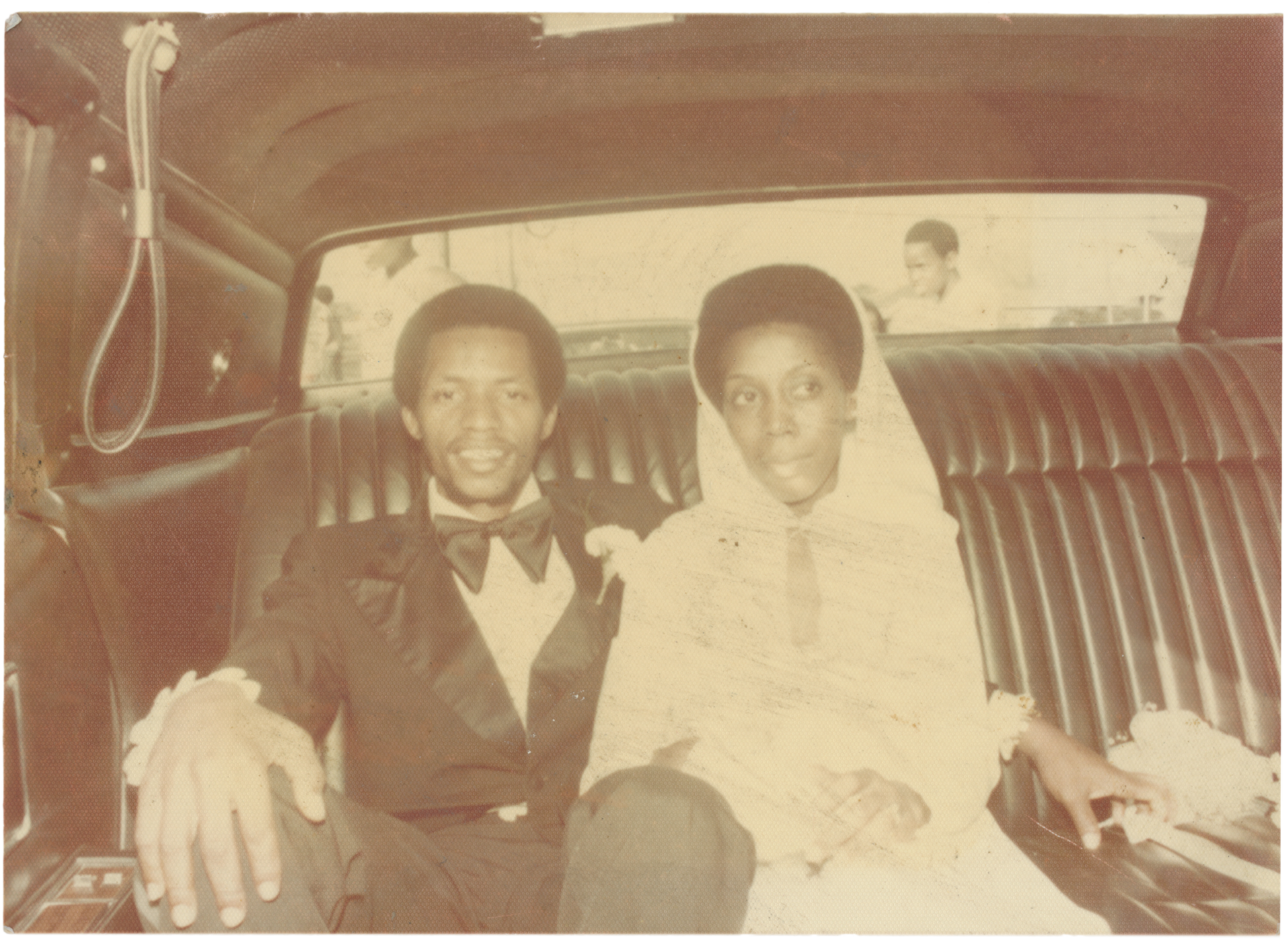 Two people seated in the back of a vintage car, smiling and dressed formally.