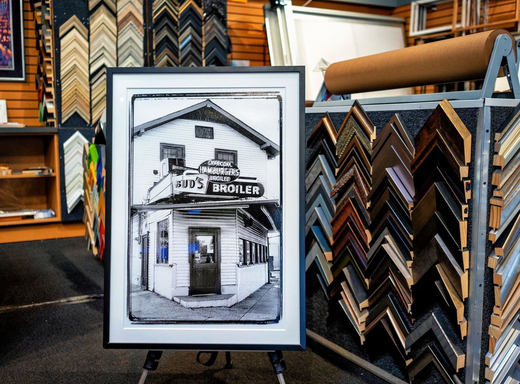 Framed black-and-white sketch of a storefront displayed in a picture frame shop surrounded by frame samples