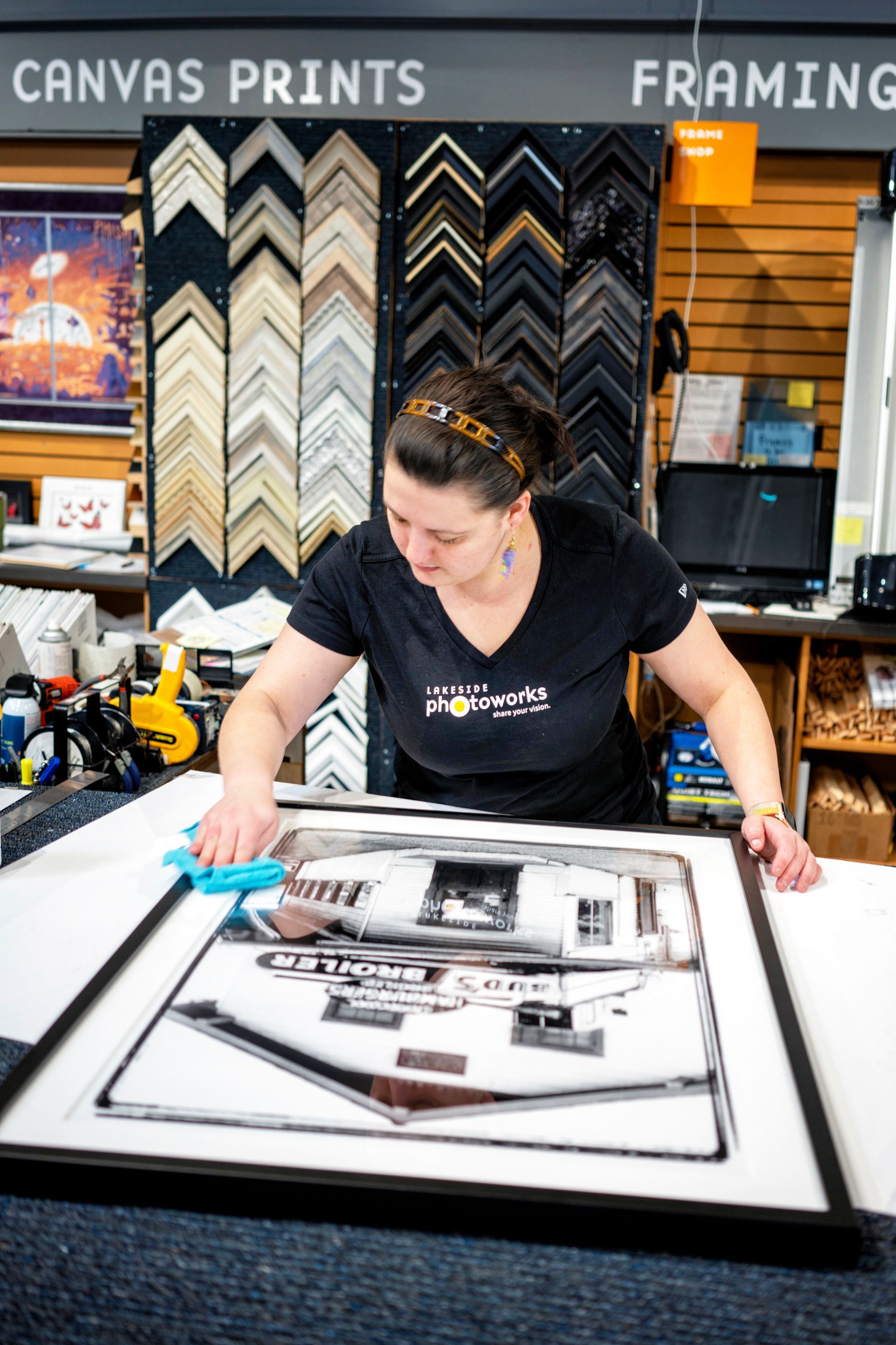 Woman assembling a framed print in a framing shop, with canvas prints displayed behind her.
