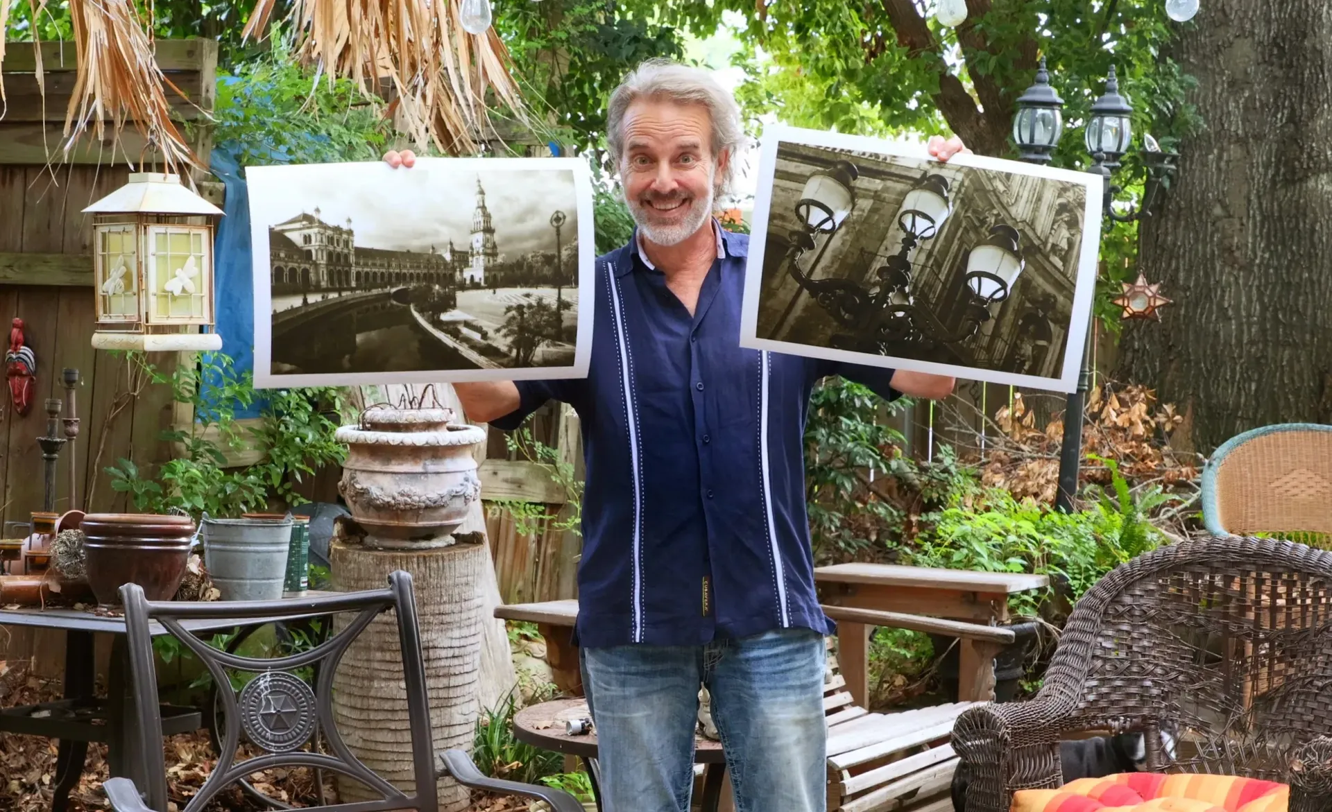 Man outdoors in a garden holding two framed black-and-white landscape photos.