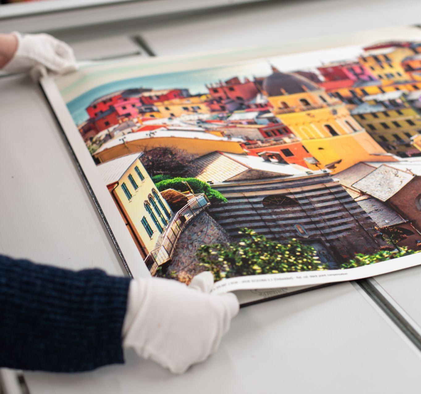 Gloved hands holding a glossy print of colorful hillside buildings on a table
