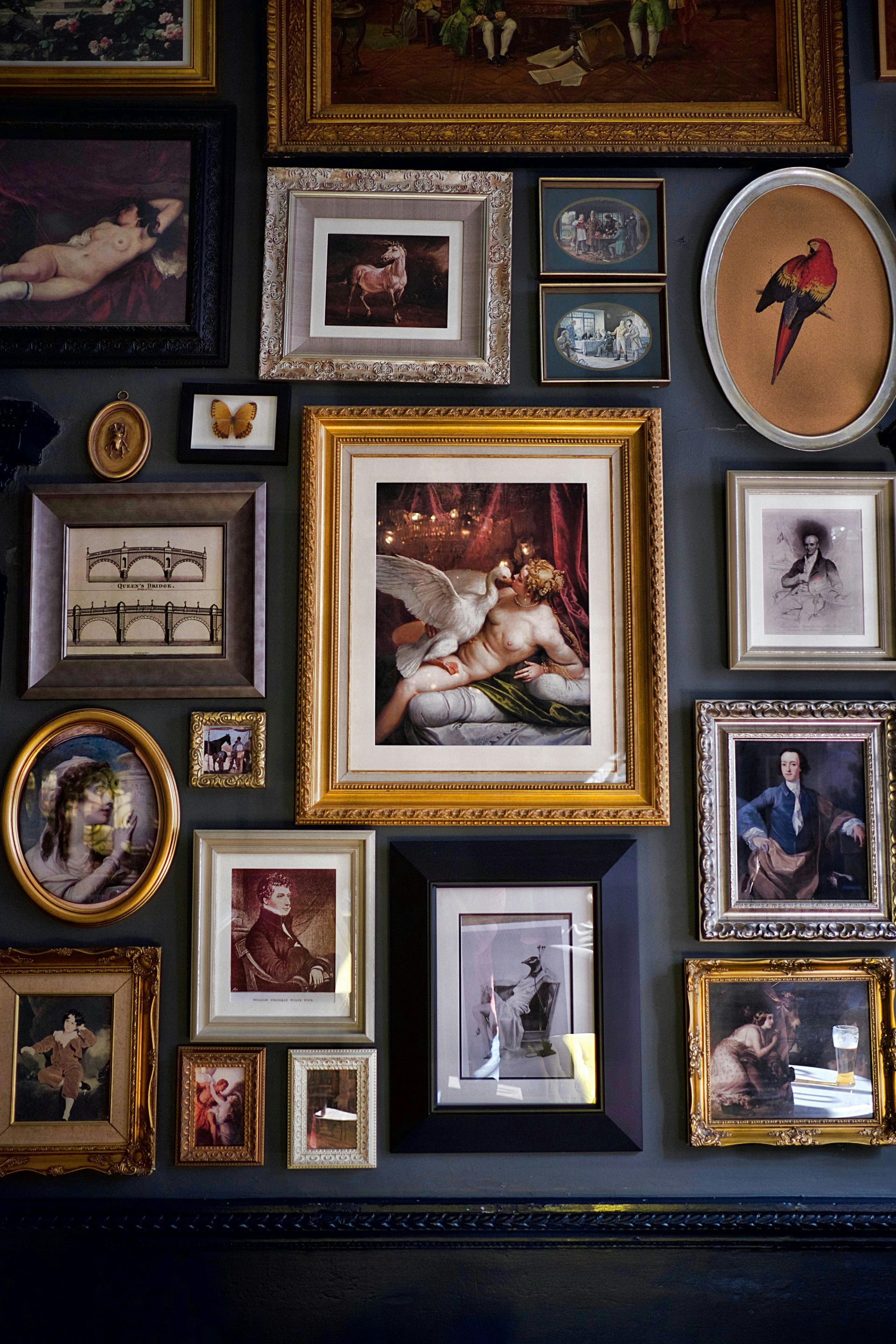 Eclectic gallery wall of framed art and photos on a dark wall, centered on a large golden-framed painting