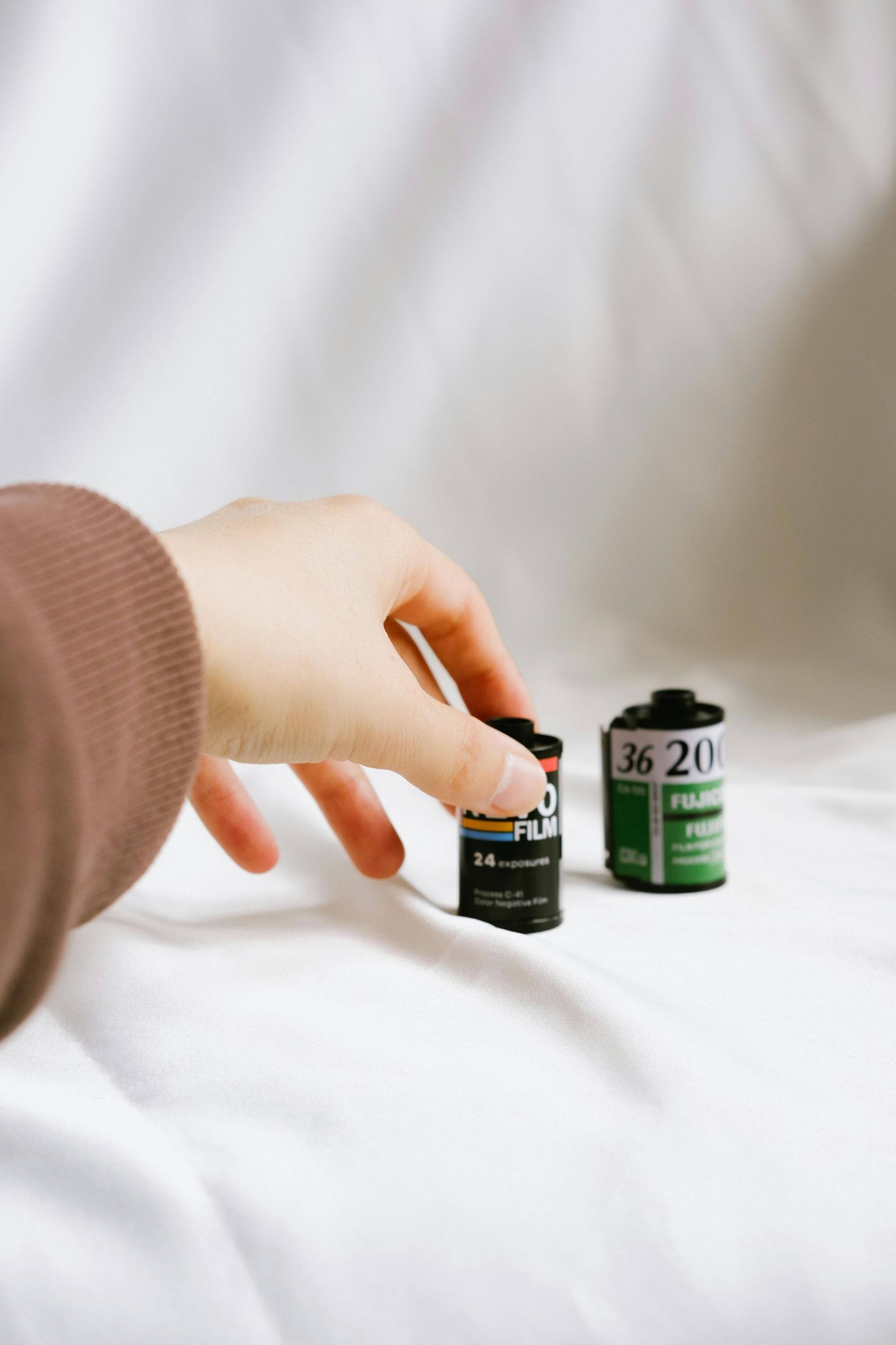 Hand reaching toward two film canisters on a white bed, one black and one green.