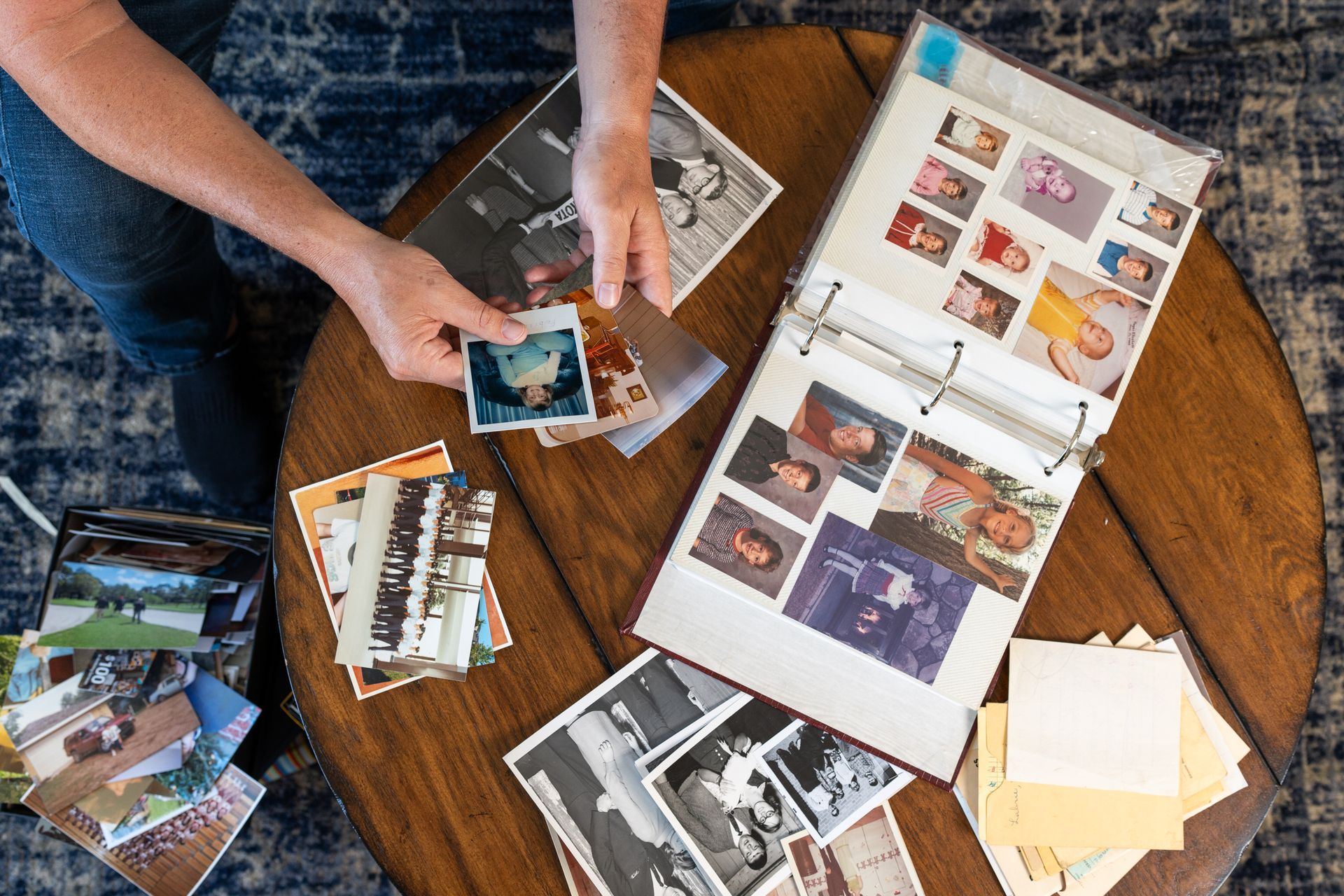 Hands arranging photo prints and an open scrapbook on a wooden table over a blue rug