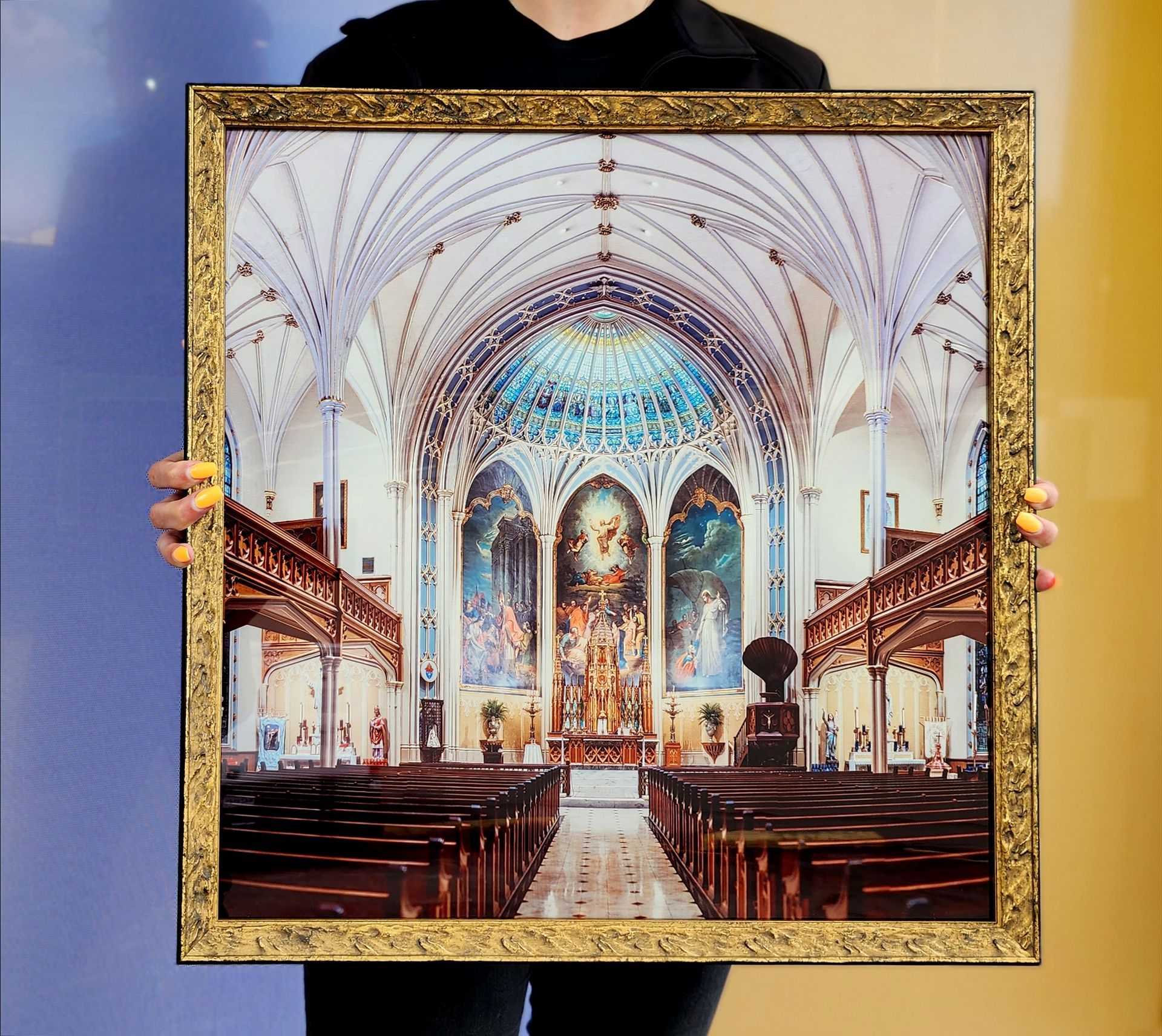 Framed painting of a bright church interior with ornate arches, blue dome, and wooden pews, held up by a person
