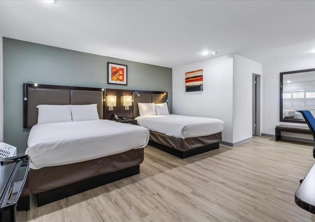 A hotel room with two queen beds featuring white bedding, brown headboards, light wood floors, and neutral walls.
