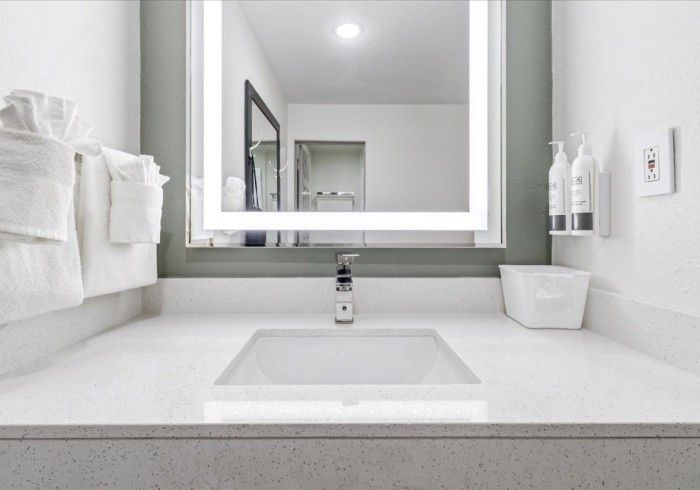 A minimalist white hotel bathroom vanity with a rectangular sink, illuminated mirror, and mounted toiletry bottles.
