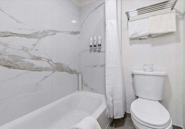 A bathroom with a white bathtub against marble-patterned tile, a white shower curtain, a white toilet, and hanging towels.