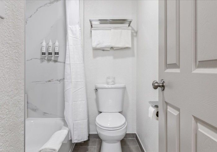 A bathroom featuring a toilet, a shower with a white curtain and marble-tiled wall, and a towel rack with white towels.
