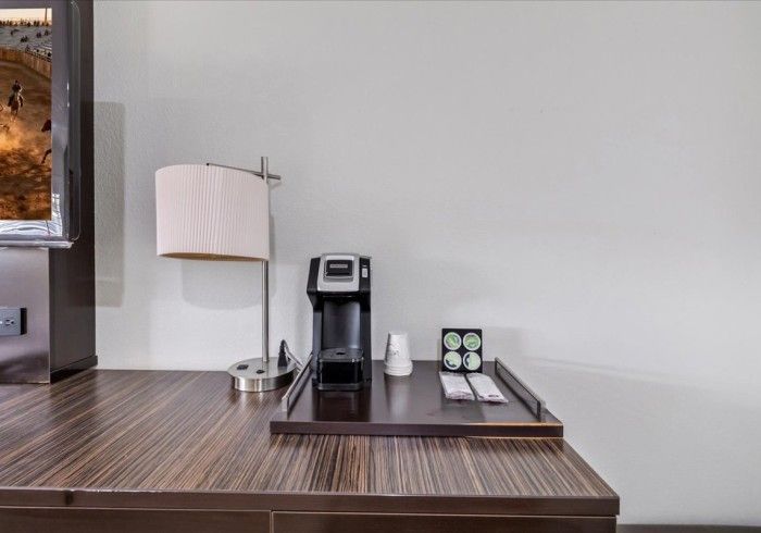 A hotel desk with a lamp, a black coffee maker, and a small tray containing coffee pods and stirrers.