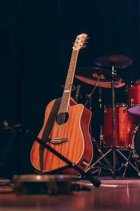 An acoustic guitar sits on a stand on a stage with a drum set partially visible in the background.