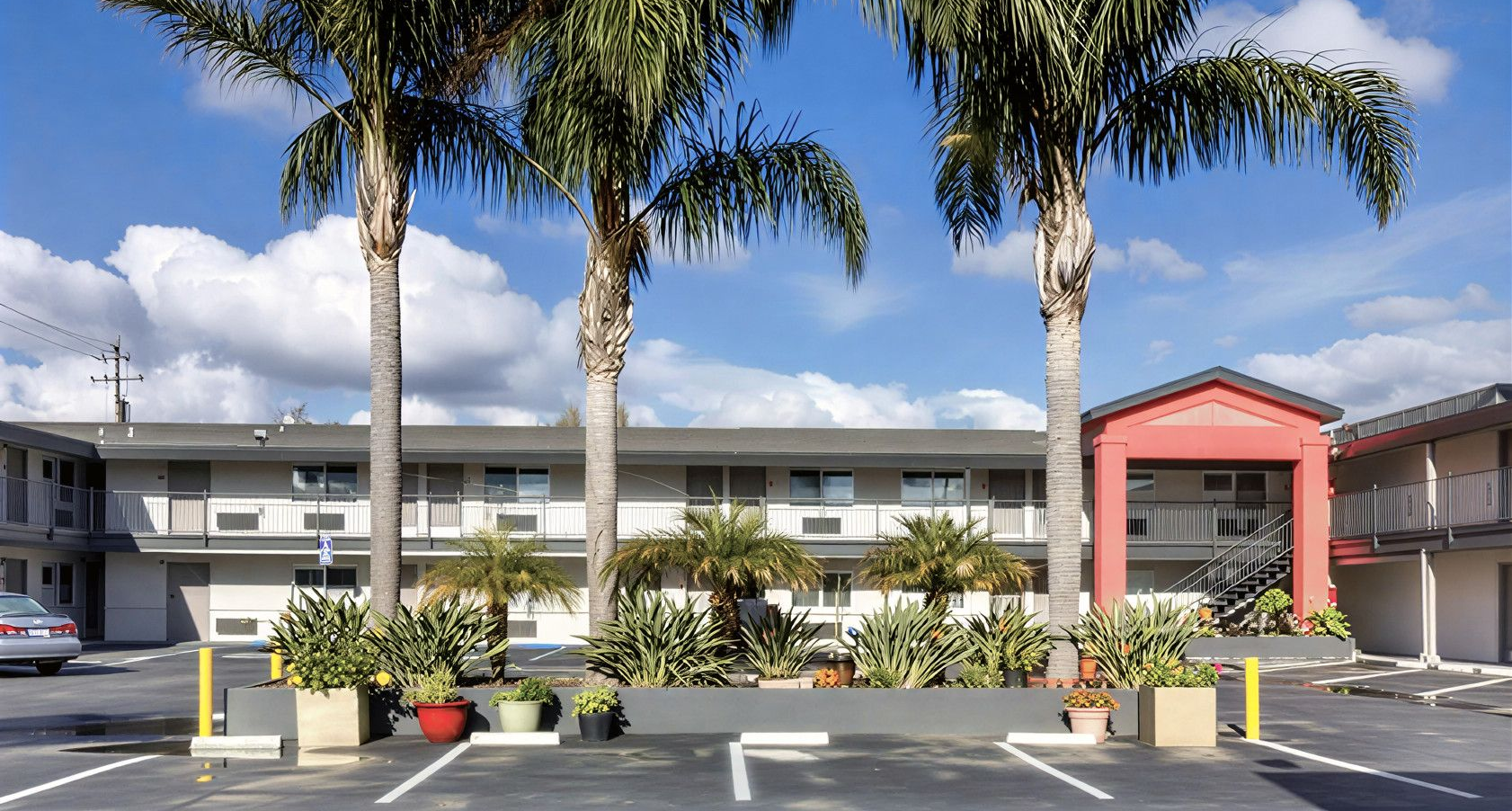 A two-story motel with a red entrance, palm trees, and potted plants in front of a parking lot under a blue sky.