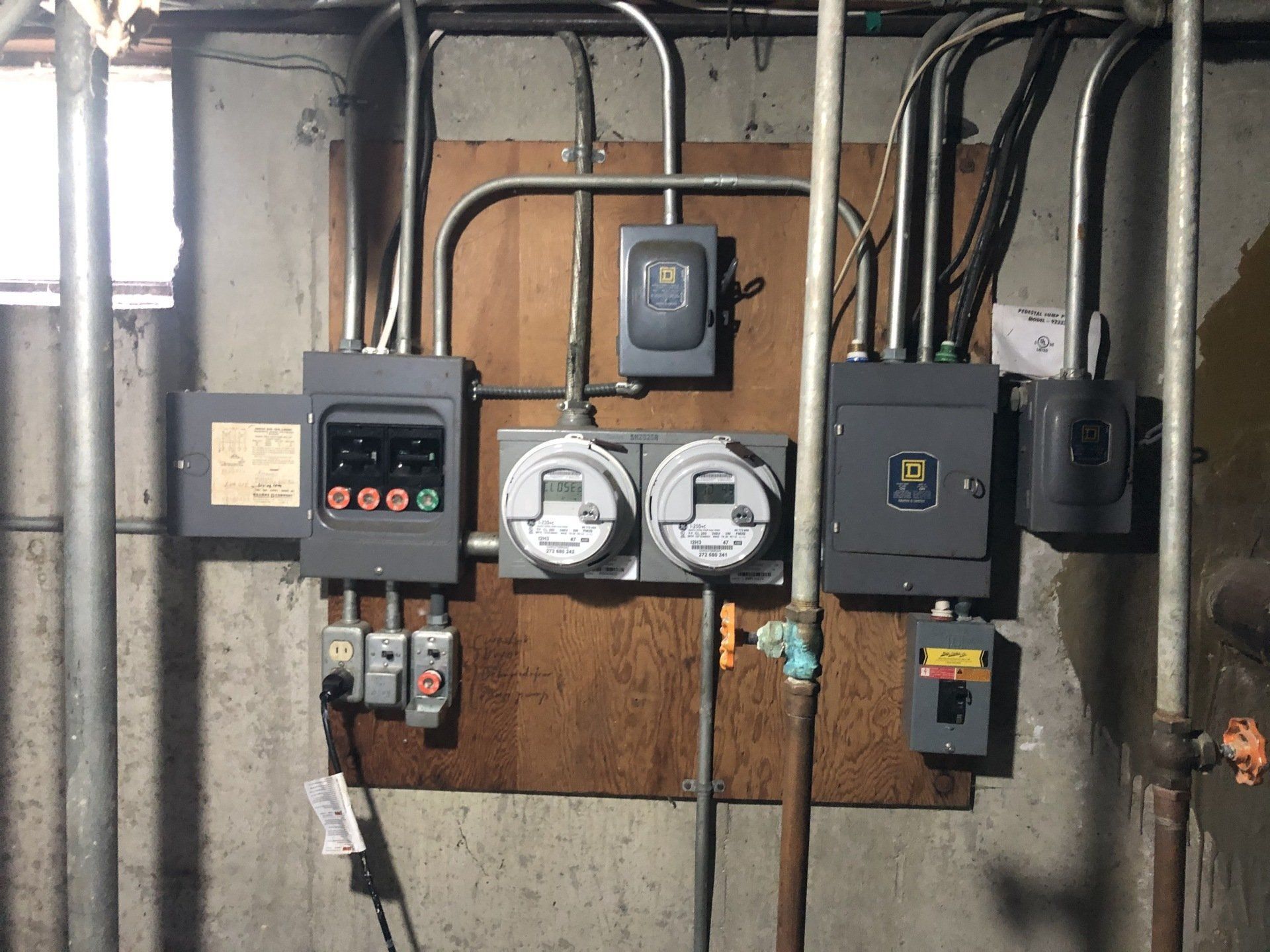 Electrical meter and breaker panel mounted on a wooden board with conduit and wiring.