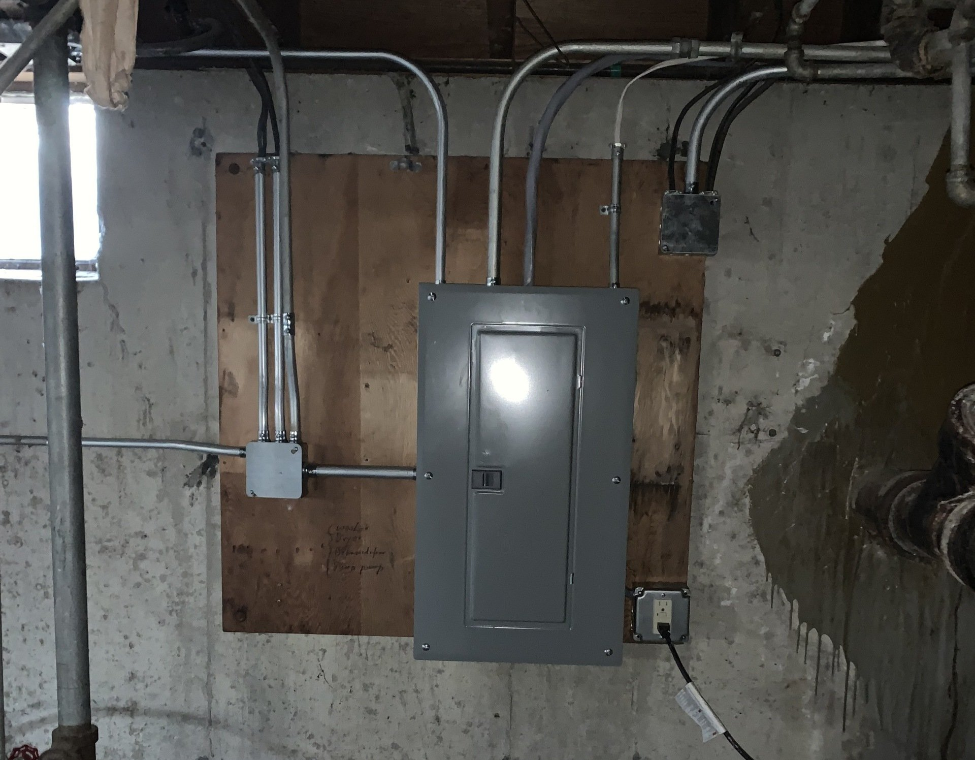 Gray electrical panel on a concrete basement wall with conduit and exposed wiring.