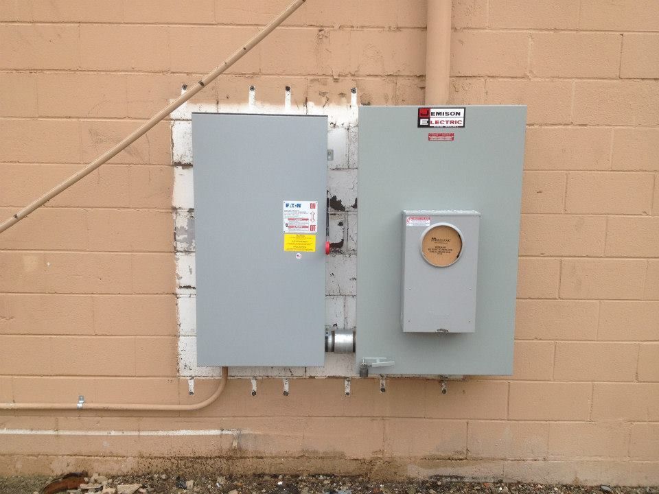Two gray electrical utility boxes mounted on a tan brick wall, with conduit and wiring visible.