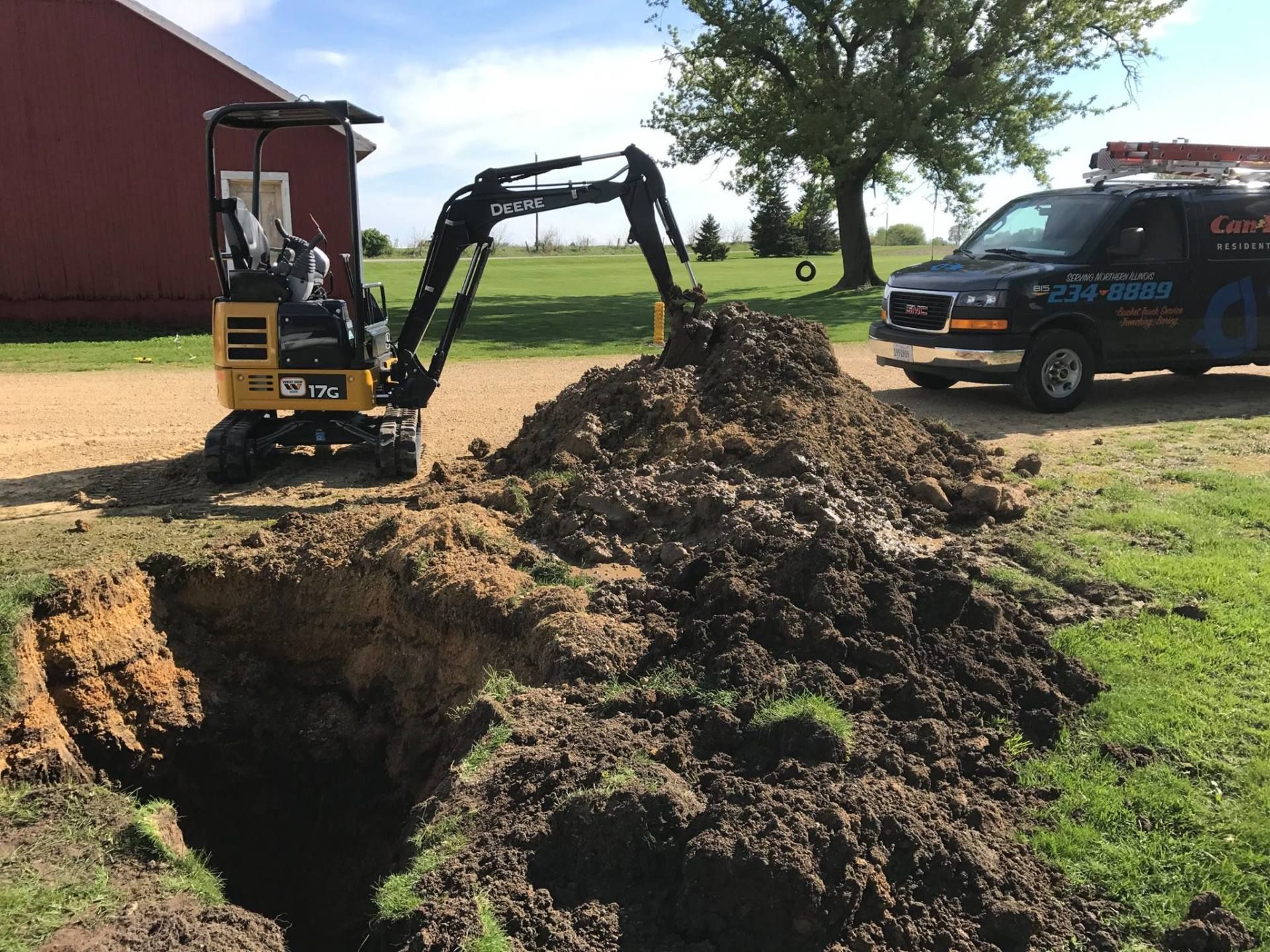 Excavator digging a large trench beside a barn, with a truck parked nearby on a grassy farm.