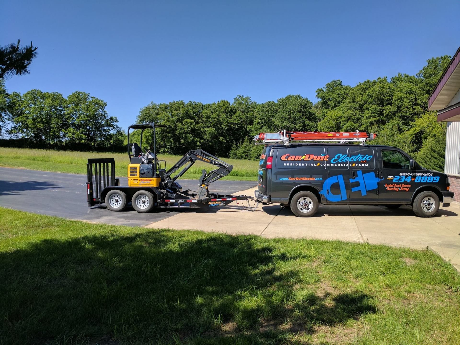 Yellow skid steer on trailer beside black service van on a sunny roadside.