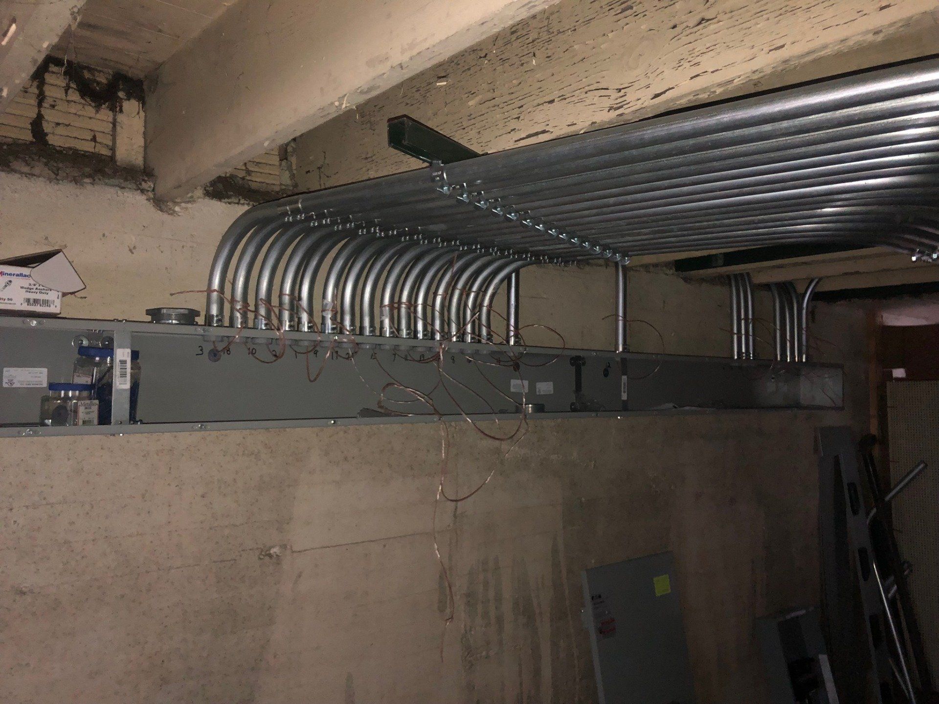 Basement utility area with metal conduit pipes running into a gray electrical panel above a concrete wall