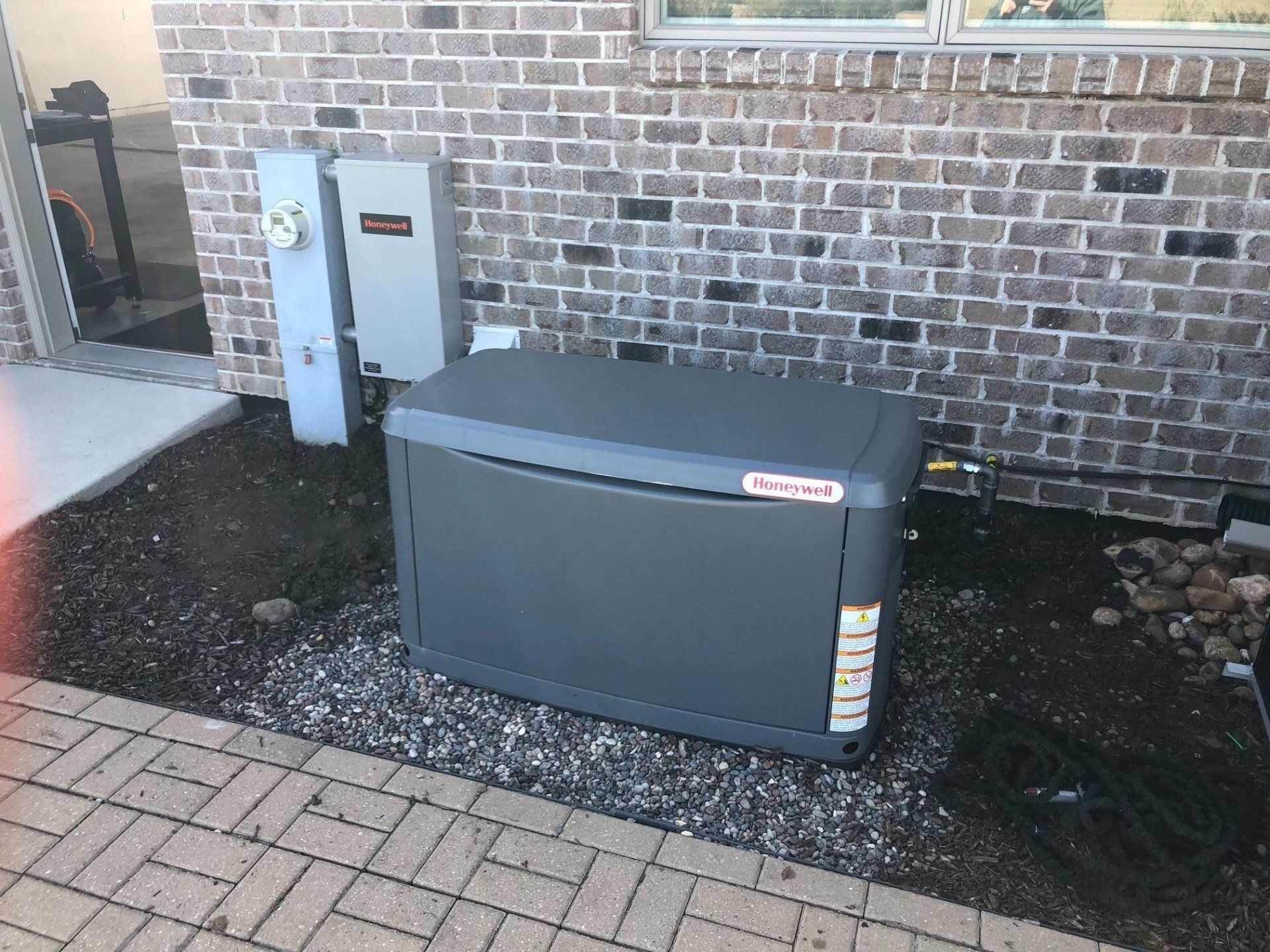 Outdoor utility area with a large gray generator beside a brick house and utility meters.