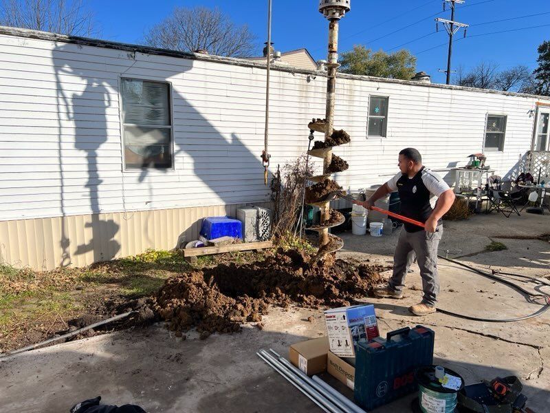 Man using a hose near a dug-up tree stump beside a white mobile home on a sunny day