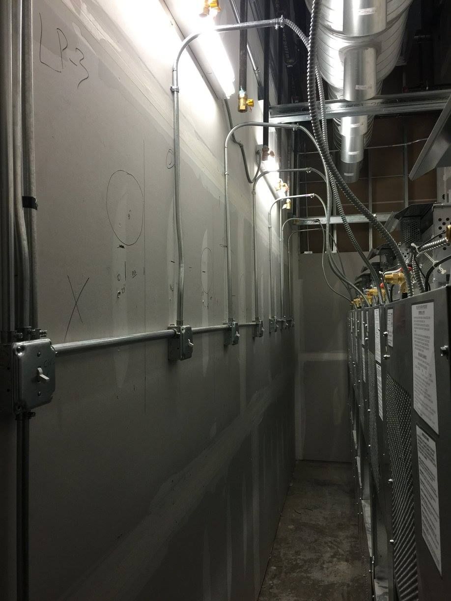 Narrow industrial hallway with metal pipes, ducts, and electrical panels along the walls and ceiling