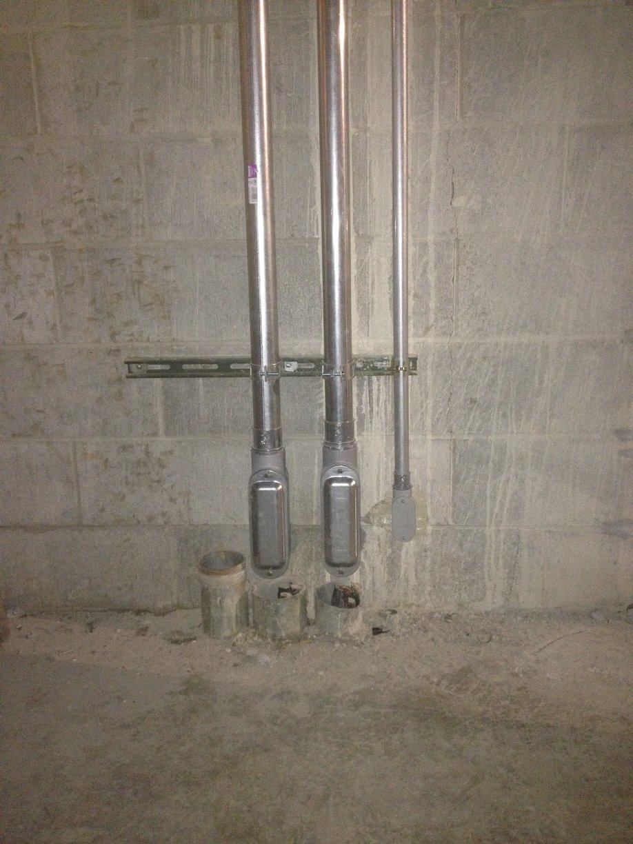 Two vertical metal pipes with gray fittings mounted on a concrete wall, with one small bucket below.