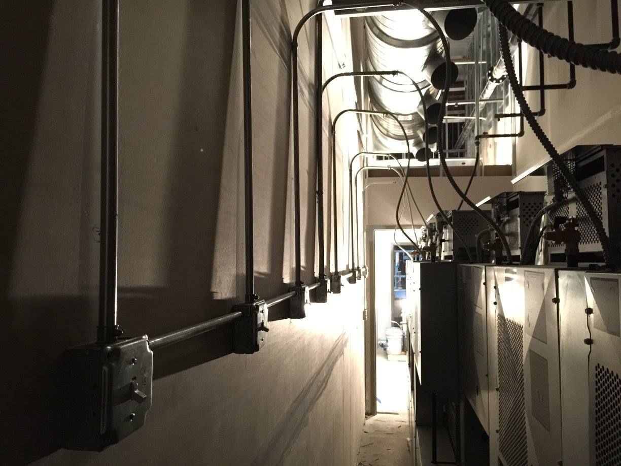 Narrow industrial hallway with exposed pipes, cables, and equipment lit by bright light at the far end