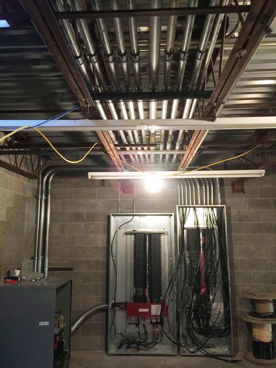 Basement utility room with exposed ceiling pipes, concrete block walls, electrical panels, and hanging cables.