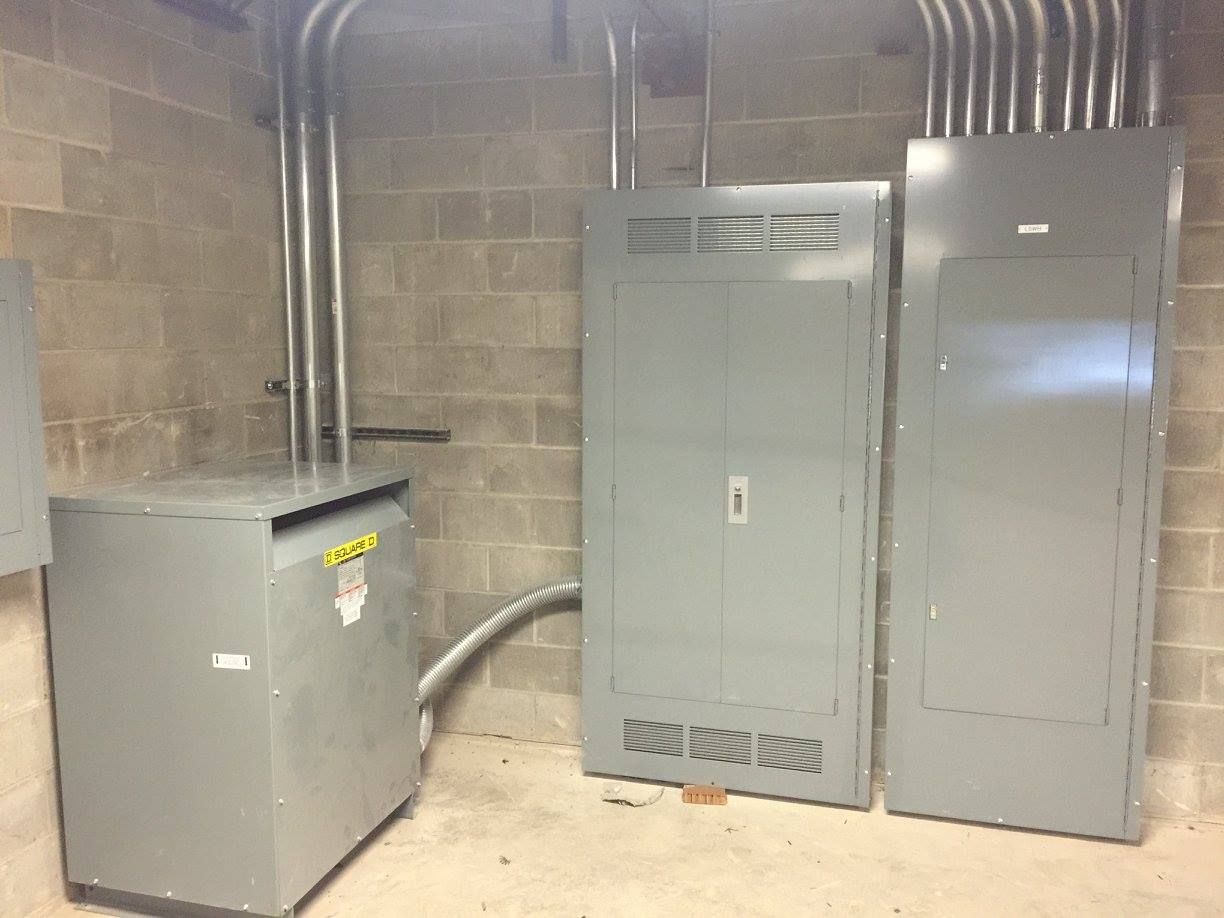 Basement utility room with electrical panels, a gray cabinet, and exposed conduit on brick walls.