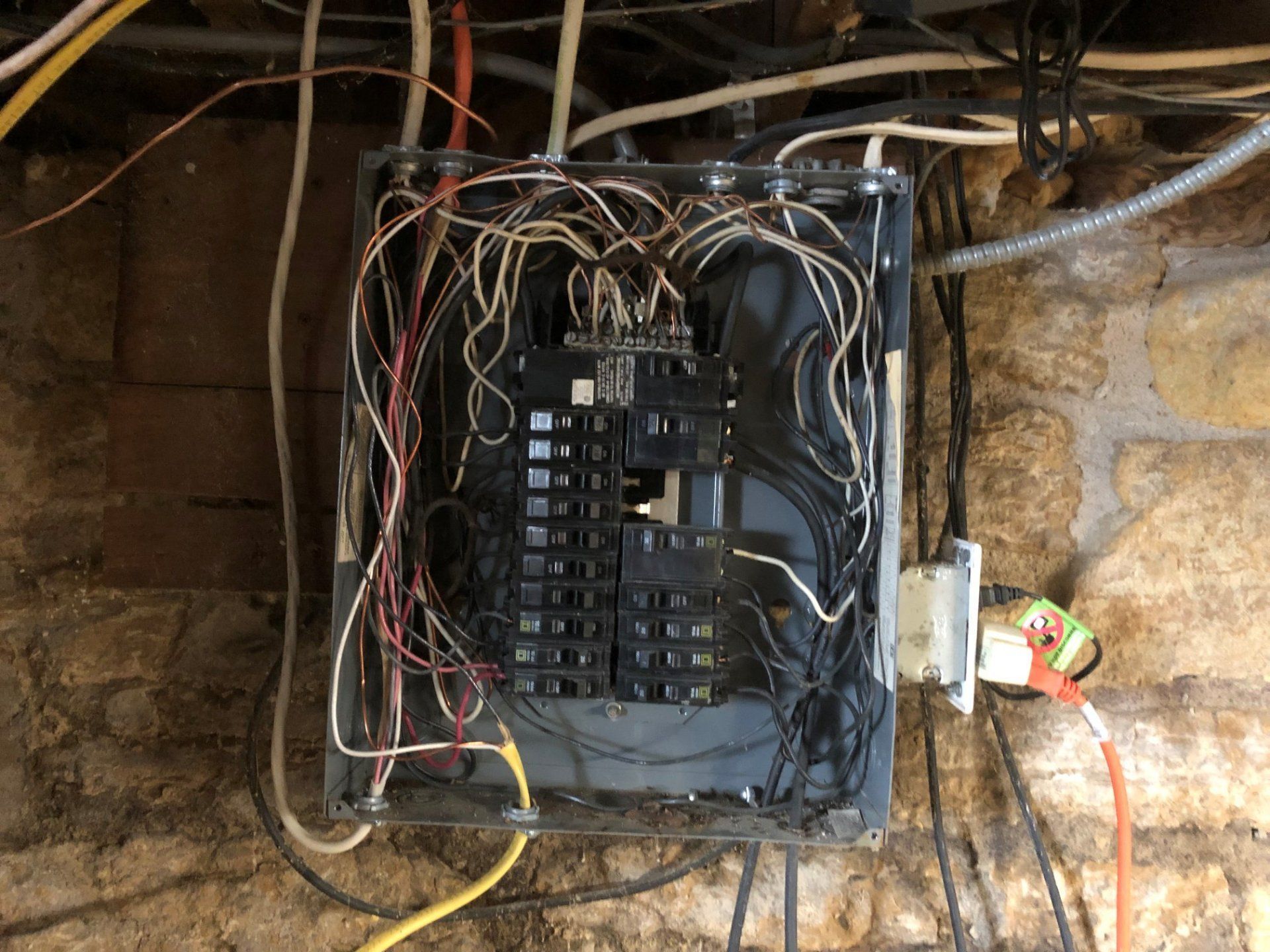 Old electrical breaker panel with tangled wires mounted on a stone wall in a dim utility area