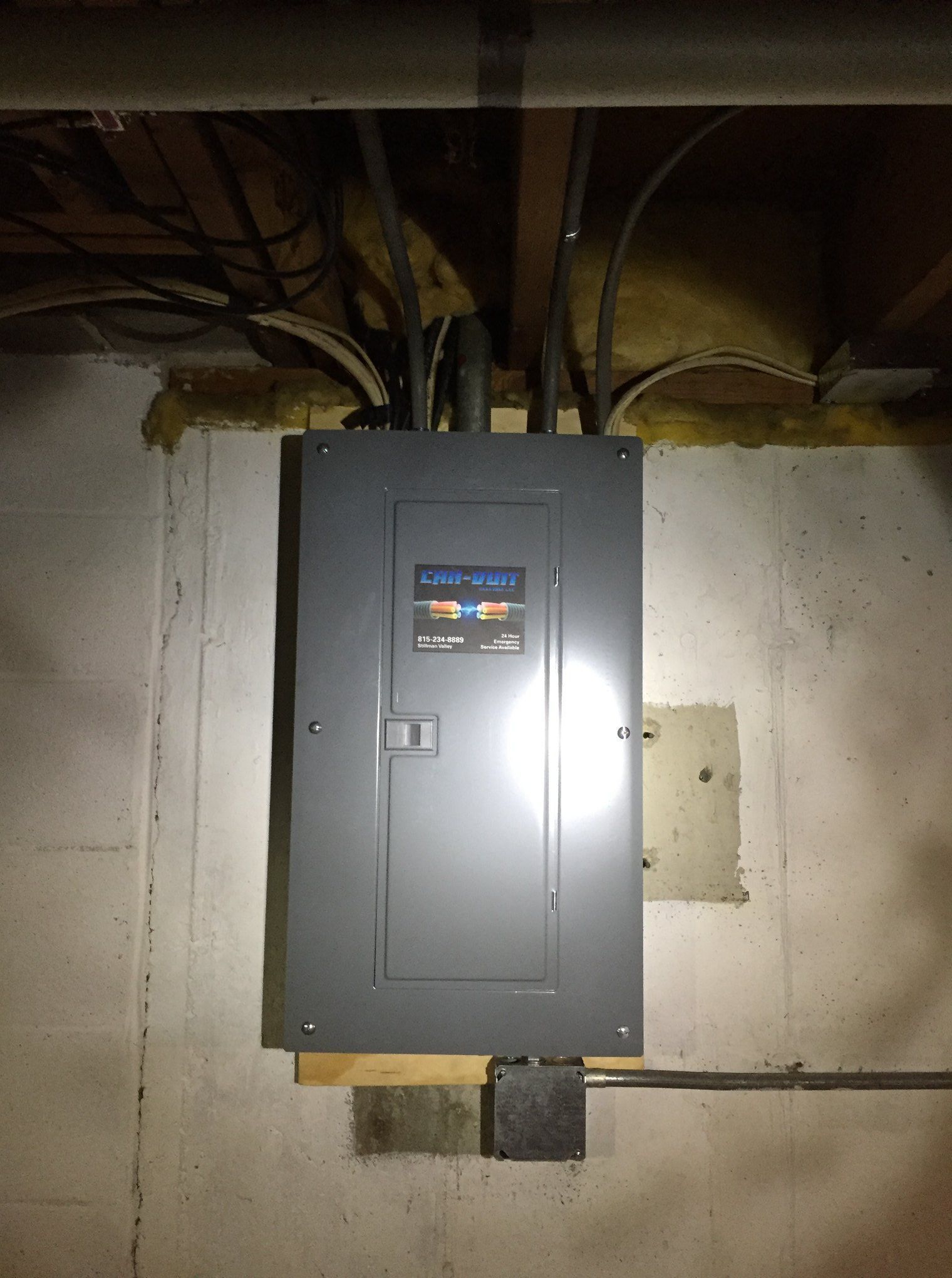Gray electrical panel mounted on a concrete wall in a basement, with exposed pipes above.