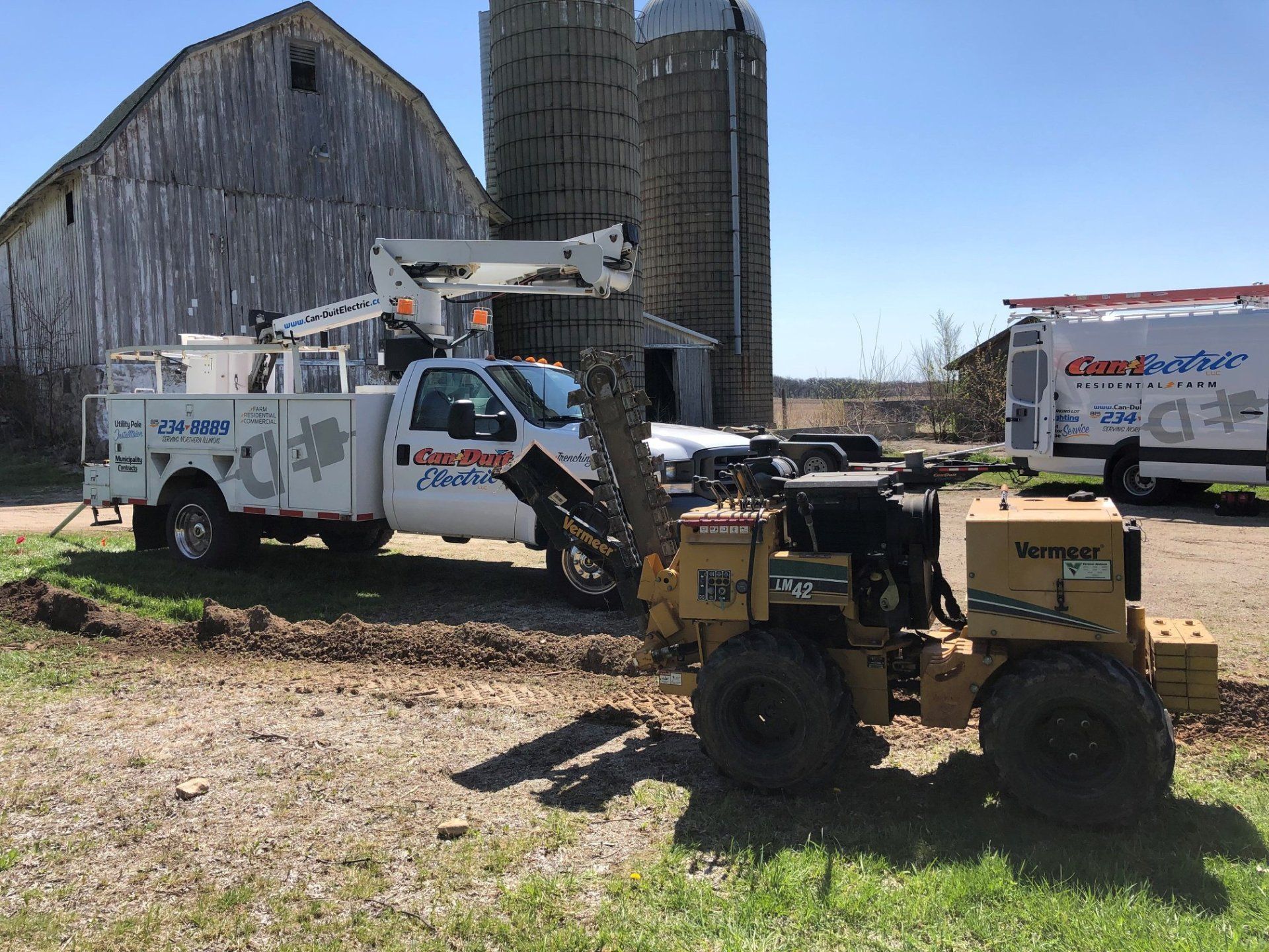 Yellow and white utility trucks and an ATV parked outside a barn and silo on a sunny farm.