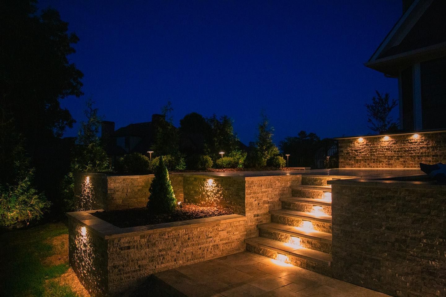 Illuminated stone steps and retaining walls in a landscaped garden at night under a deep blue sky
